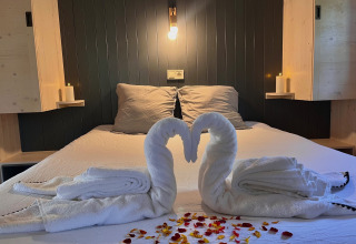 Cama romántica con cisnes de toalla, velas y pétalos de rosa en Charme hut con jacuzzi, Camping Le Périgord, Francia.