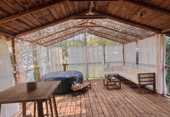 Terraza de madera cubierta con jacuzzi, zona de asientos y cortinas en Charme hut met jacuzzi, Camping Le Périgord.