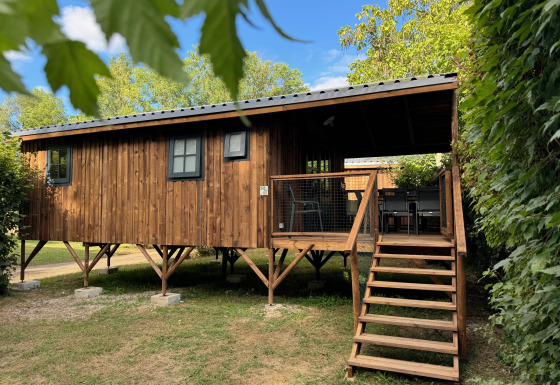 Cabaña Premium Hut de madera sobre pilotes, con terraza y escaleras, rodeada de árboles y cielo azul.
