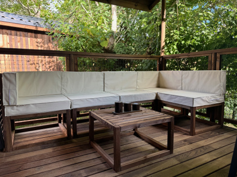 Zona de estar exterior con muebles de madera y cojines blancos en Premium Hut en Camping Le Périgord, Francia.
