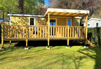 Træhytte kaldet Classic Cottage med stor terrasse og markise omgivet af grønne træer og græsplæne.