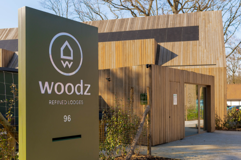 Toegang tot Woodz refined lodges, met een modern houten bord en gebouw omringd door natuur.
