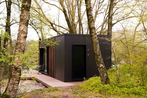 Das schwarze Nokken NKN-18 Tiny House steht in einem bewaldeten Gebiet, umgeben von Bäumen und Grün.