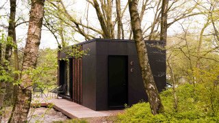 Black Nokken NKN-18 tiny house cabin with a wooden deck, nestled among trees and greenery in a forest.