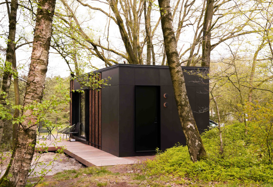 Zwarte Nokken NKN-18 tiny house met houten terras, omringd door bomen en groen in een bosgebied.