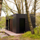 Sort Nokken NKN-18 tiny house i en skov med træer og grønne omgivelser, træterrasse foran døren.