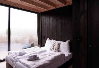 Dormitorio moderno en Nokken NKN-18 Cabin, Wilsumer Berge, Alemania, con paredes de madera oscura y grandes ventanales.