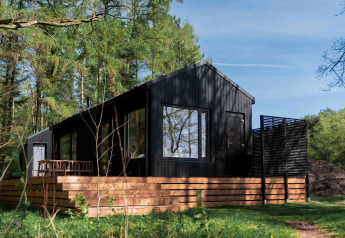Sort træhytte kaldet Koya Tiny Cabin med terrasse, omgivet af skov i Wilsumer Berge, Tyskland.