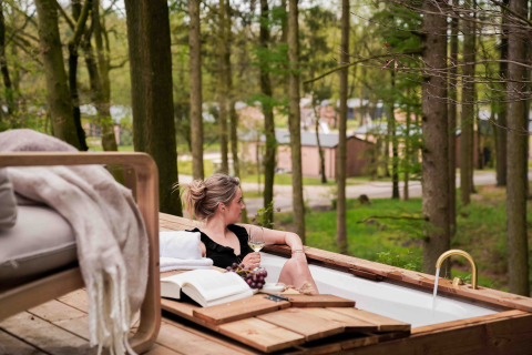 Mujer relajándose con vino en una bañera exterior en Koya Tiny Cabin, rodeada de bosque en Wilsumer Berge.