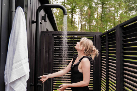 Mujer disfrutando de una ducha exterior en Koya Tiny Cabin en Wilsumer Berge, Alemania, rodeada de madera oscura