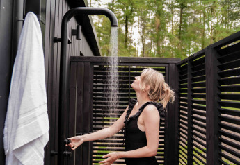 Mujer disfrutando de una ducha exterior en Koya Tiny Cabin en Wilsumer Berge, Alemania, rodeada de madera oscura