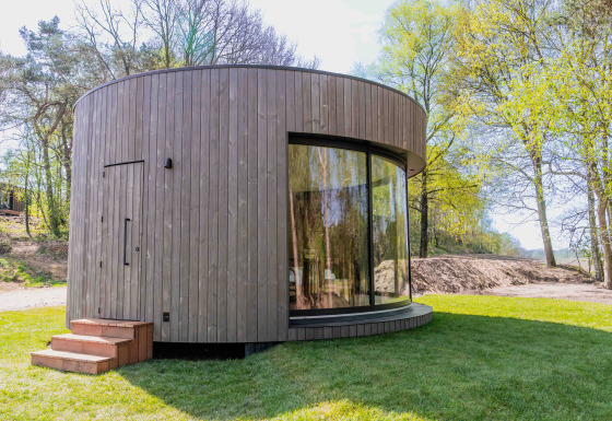 Casa diminuta redonda llamada Lumni Pod con fachada de madera y grandes ventanales en entorno natural verde.