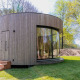 Moderne, rund tiny house Lumni Pod med træbeklædning og store vinduer i naturskøn grøn omgivelser.