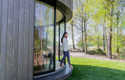Une femme se tient devant la tiny house Lumni Pod à Wilsumer Berge, Allemagne, entourée de verdure et d'arbres.