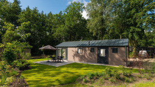 Velthorst Comfort Lodge mit grüner Rasen, Gartentisch, Stühlen, Sonnenschirm, umgeben von Bäumen.