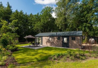 Velthorst Comfort Lodge mit grüner Rasen, Gartentisch, Stühlen, Sonnenschirm, umgeben von Bäumen.