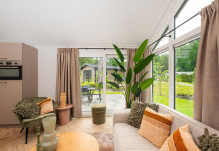 Stue i Velthorst Comfort-lodge på Veluwepark de Bosgraaf, med store vinduer, planter og hyggelig indretning.