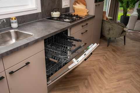 Modern kitchen inside a Dutch lodge with an open dishwasher, gas stove, and living area in the background.