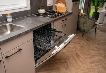 Cocina moderna en una cabaña holandesa con lavavajillas abierto, estufa de gas y sala de fondo.
