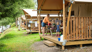 Familie nyder tid sammen på verandaen af et safaritelt omgivet af natur og træer i en campingplads.