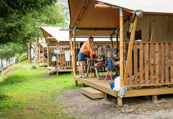 Familie nyder tid sammen på verandaen af et safaritelt omgivet af natur og træer i en campingplads.