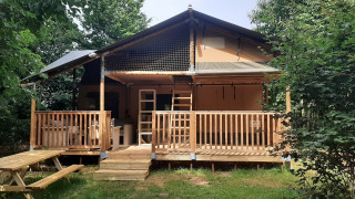 Tienda safari llamada Ranger Lodge con porche de madera, rodeada de árboles y banco de picnic adelante.