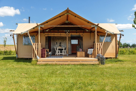 Safaritent met overdekte veranda op een grasveld onder een blauwe hemel, voorzien van comfortabel meubilair.