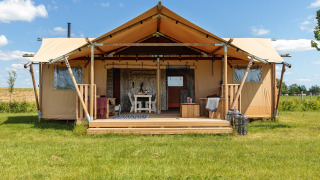 Safaritent met overdekte veranda op een grasveld onder een blauwe hemel, voorzien van comfortabel meubilair.