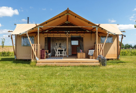 Safaritent met overdekte veranda op een grasveld onder een blauwe hemel, voorzien van comfortabel meubilair.
