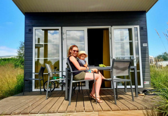 Donna e bambino seduti fuori da una tiny house Comfort Lodge al Kompas Camping Westende in Belgio.