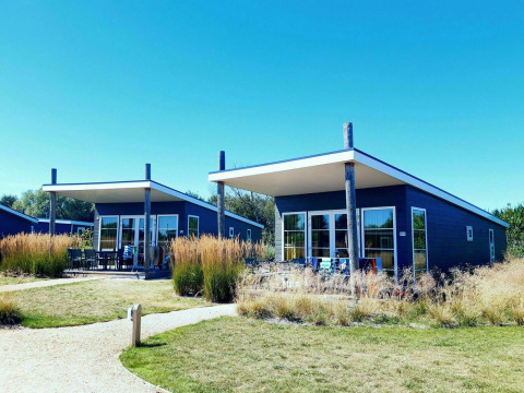 Twee moderne blauwe lodges met terras, omgeven door gras en wandelpaden bij Kompas Camping Westende, België.