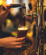 Persona sirviendo cerveza de barril en un bar con ambiente de parque vacacional y glamping al fondo.