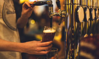 Persona sirviendo cerveza de barril en un bar con ambiente de parque vacacional y glamping al fondo.