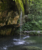 Small waterfall trickling from mossy rock into a tranquil pond, surrounded by greenery at a holiday park.