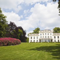 Weißes Herrenhaus, umgeben von grünen Gärten und Blumen, in einem Ferienpark mit Glamping-Angeboten.