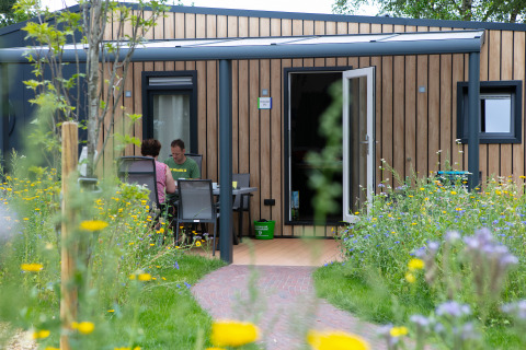To personer sidder ved et bord foran et moderne træhus omringet af vilde blomster i en feriepark