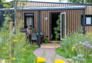 To personer sidder ved et bord foran et moderne træhus omringet af vilde blomster i en feriepark