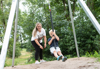 Kvinde leger med barn på svævebane i Holiday Park 't Rheezerwold omgivet af grønne træer i Overijssel.