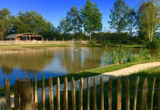 Blick auf einen ruhigen Teich mit Springbrunnen, Bäumen und einer Holzhütte im Holiday Park ’t Rheezerwold.