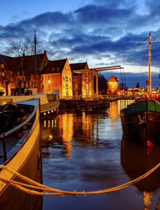 Vista nocturna de barcos amarrados, paseo empedrado y luces festivas en parque de glamping.