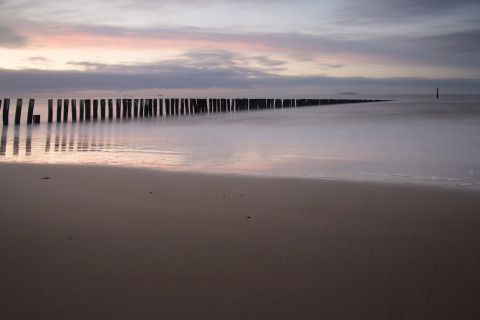 Ruhiger Strand bei Sonnenuntergang nahe Tholen, Zeeland, Niederlande, mit Pfählen im Wasser und sanften Farben.