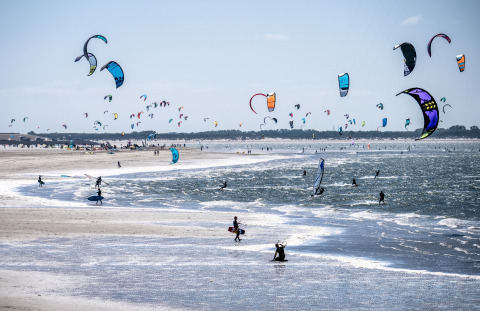 Strand fyldt med farverige drager og glade mennesker, der kitesurfer ved en feriepark med glampingmuligheder.