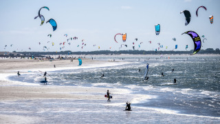 Cielo lleno de cometas mientras personas hacen kitesurf en la playa junto a un parque de vacaciones glamping.