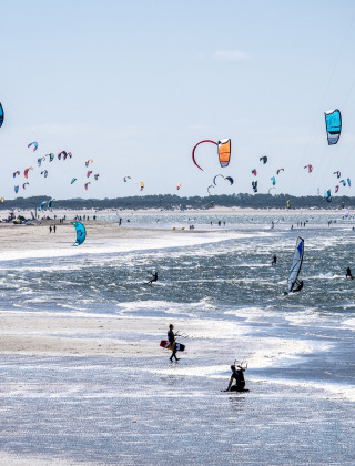 Cielo lleno de cometas mientras personas hacen kitesurf en la playa junto a un parque de vacaciones glamping.