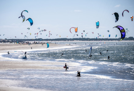 Kites en kitesurfers op een druk strand bij een vakantiepark met glamping-accommodaties aan zee.