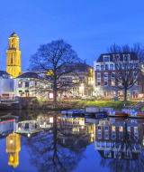 Vista nocturna de una ciudad junto al río, barcas, alojamientos glamping y torre iluminada en parque vacacional.
