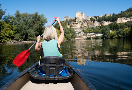 Persona remando en canoa por un río con vista a un castillo en la colina, rodeado de árboles y cielo azul.
