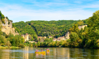 Folk padler i kano på en flod ved en feriepark omgivet af skovklædte bakker og charmerende bygninger.