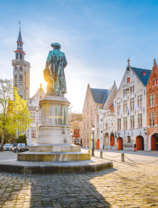 Plaza histórica de Bélgica con casas de colores, calles adoquinadas y una estatua central al sol.