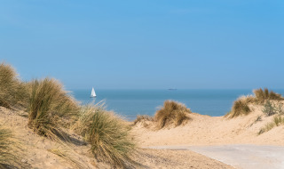 Zandduinen en zeezicht nabij Westende, België, met helmgras en een zeilboot aan de horizon.